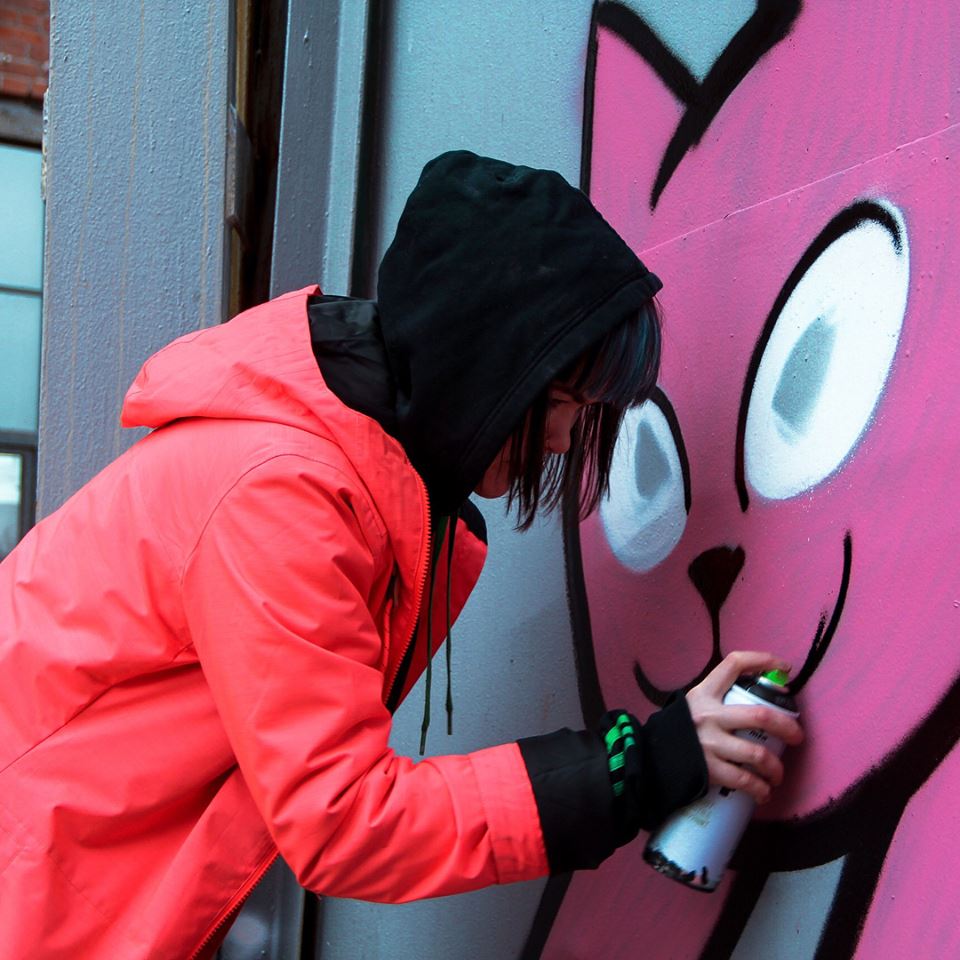 woman in orange coat spraypaints a large pink cat onto a wall